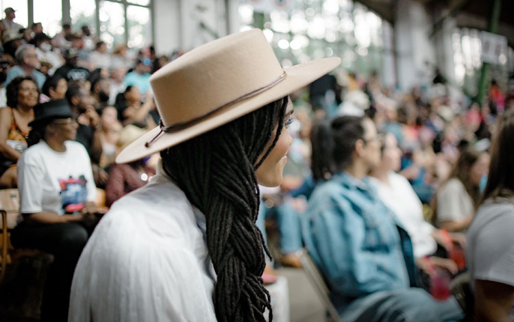 A woman sitting in a crowd at a rodeo looking away from the camera wearing a tan felt hat